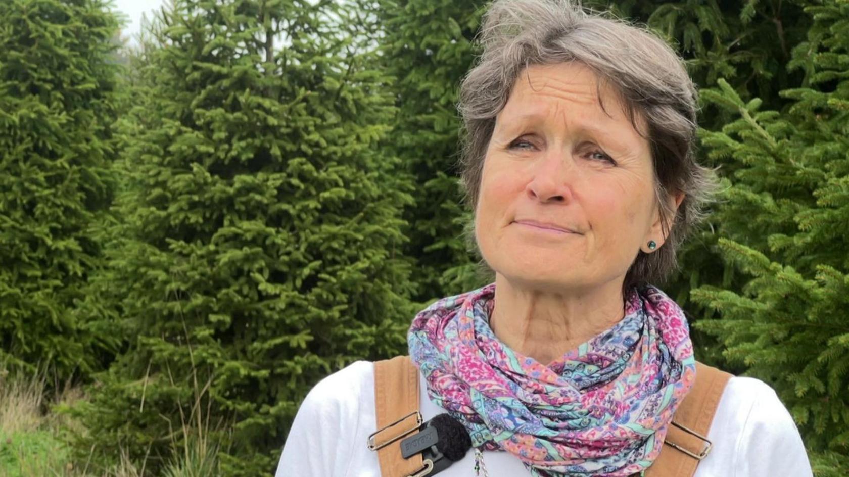Jane Ingram, wearing a white top and pink and turquoise patterned scarf, standing in front of a plantation of mature Christmas trees.