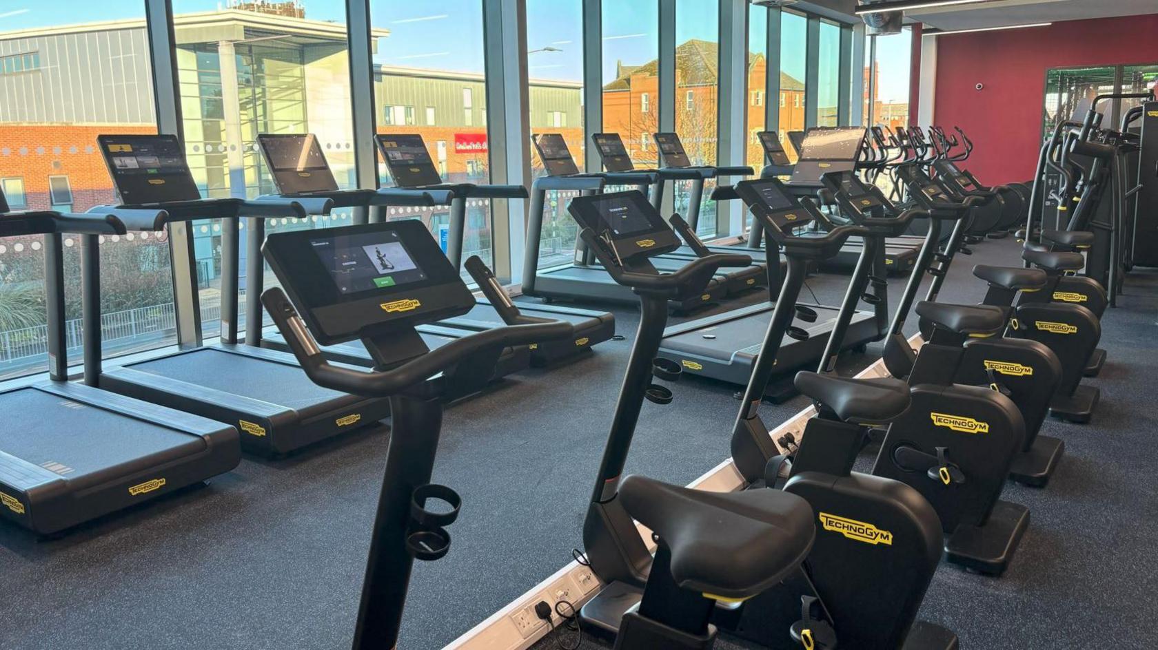 Running machines and five black exercise bikes visible on the gym floor. One side of the gym is floor to ceiling height windows which overlook a street.