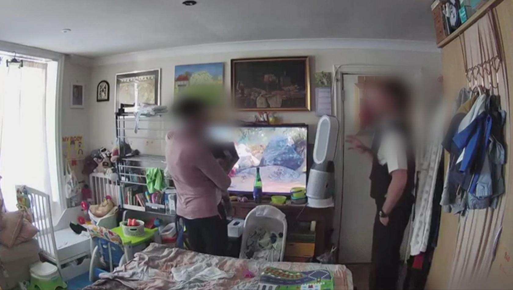 A still from home security footage showing a woman talking to a police officer in a crowded room with a TV by the door, a clothes drying rack propped up in front of some shelves, a cot in one corner, a fan by the TV, coats hung up on one wall and what appears to be the bed in the middle. There is barely enough space for the adults to stand up.