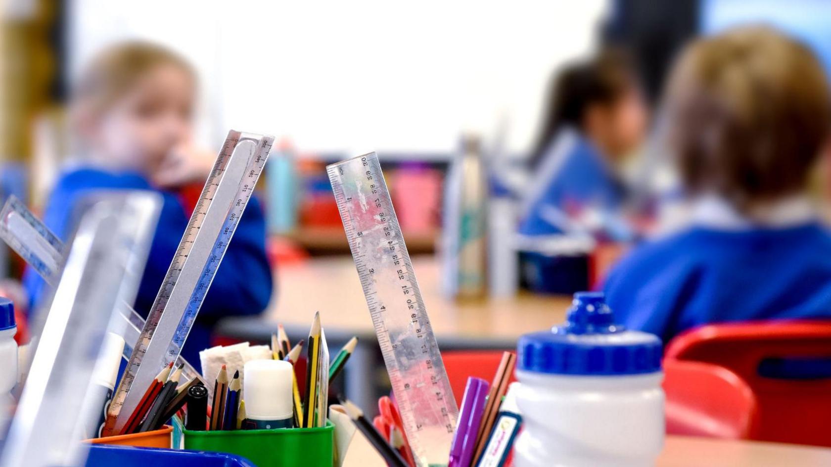 Children sat in a classroom who are blurred out and can't be identified with rulers, pencils and super-glue in pots in the foreground.
