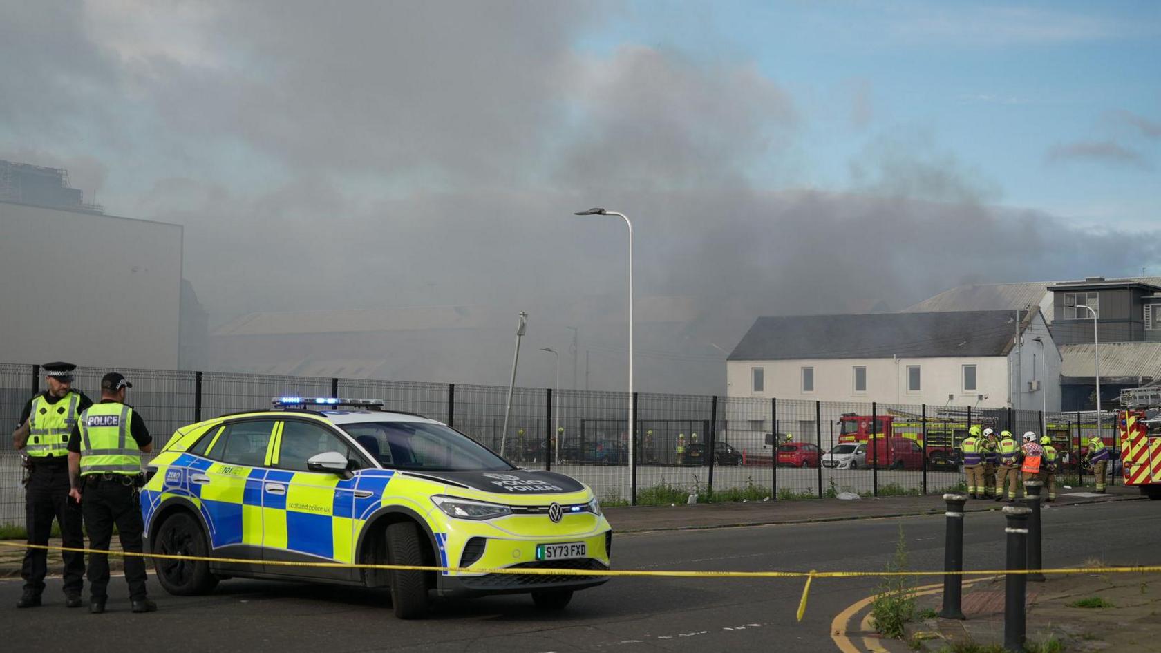 Police stand by a cordoned off road with a police car behind them. Lots of grey smoke billows in the distance and fire crews can be seen further down the road by fire engines.