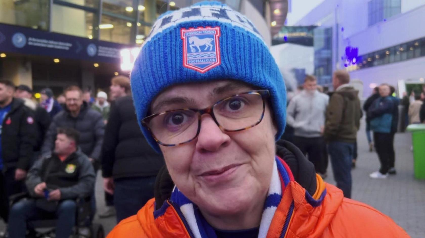 A woman is wearing glasses and her hair is hidden by an Ipswich Town woolly hat.  It's blue and white with an ITFC badge in the centre. She is wearing a bright orange coat with an ITFC scarf.  In the background there are more supporters and the outside of the Tottenham Hotspur Stadium.