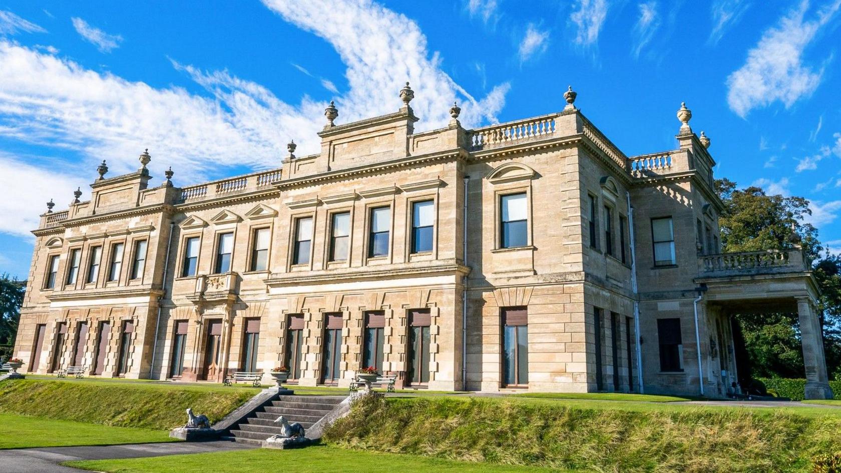 The front and side of a large stone built house with a parapet on the roof, decorated with vase shaped pediments. The house is on a raised area of grass with a stone staircase leading down to a lower level of grass with a path running off to the left.