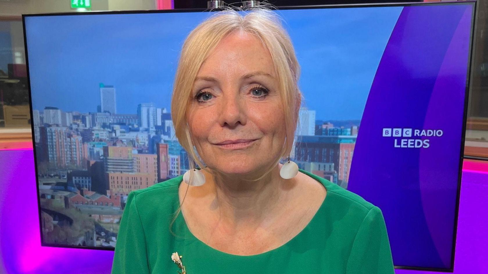 The Mayor of West Yorkshire, Tracy Brabin, in the centre of the frame. She is wearing a green dress. Behind her is a screen with a view of the Leeds skyline and a BBC Radio Leeds logo on the right-hand side.