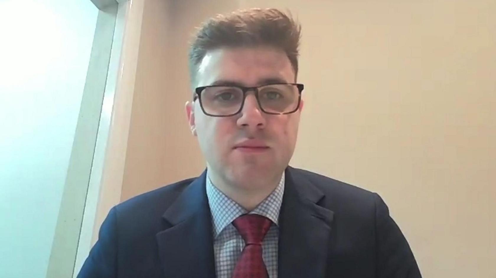 DUP councillor Dean McCullough - he has short brown hair and black-rimmed glasses and is wearing a dark suit jacket over a white-and-blue checked shirt with a maroon tie.