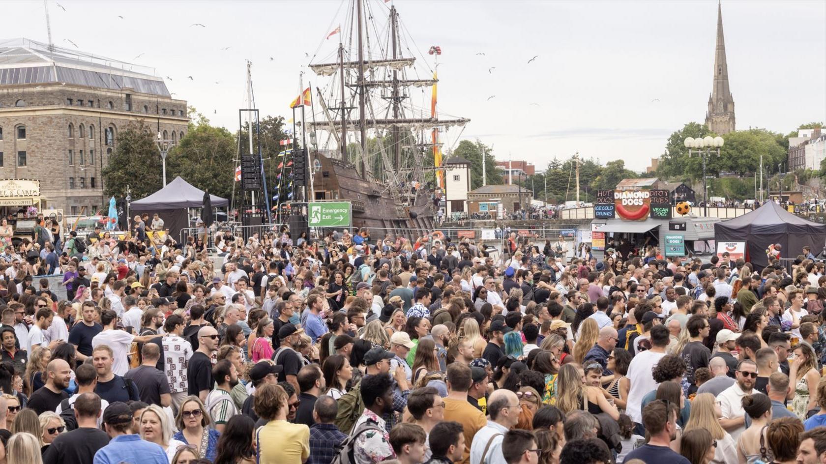 A large crowd of people standing in Lloyd's Amphitheatre during Bristol Harbour Festival. The sky is overcast but bright, and in the background you can see a tall galleon ship moored in the harbour, with Spanish flags and bunting flying from its mast.