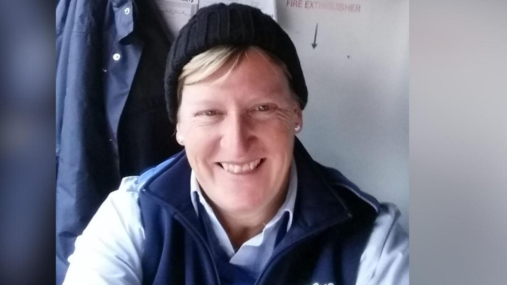 A woman with blonde hair poking out from under a black beanie hat grins. She is sitting down and wearing a blue shirt and navy bodywarmer.