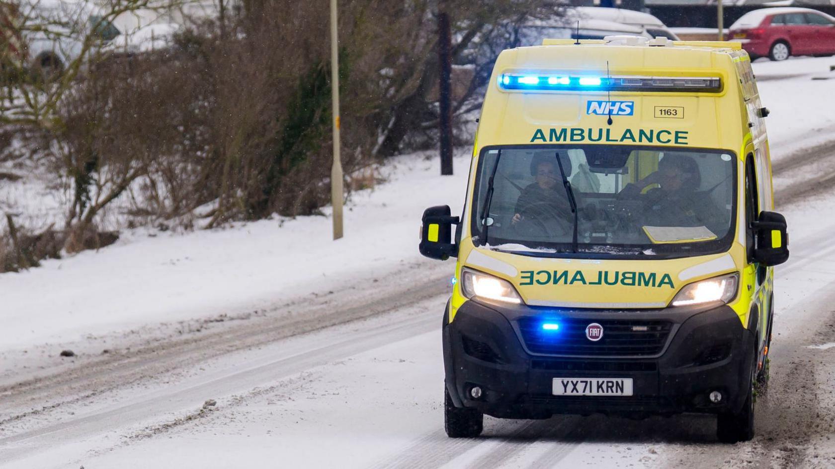 A yellow ambulance with its blue emergency lights on is driving along a snowy road. The grey tarmac is only peeking through where other cars have driven along it.