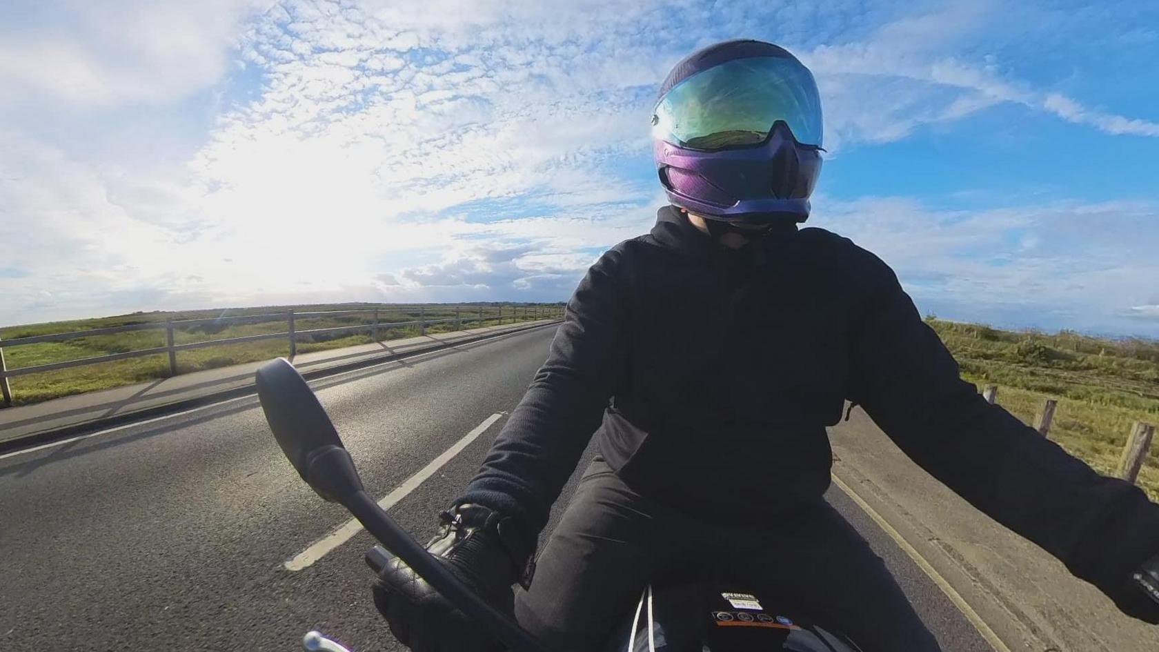 Sam Arnett riding his motorcycle on a sunny day. He has a purple helmet on and a black jacket with gloves. He is on a country road which has grass either side of it.