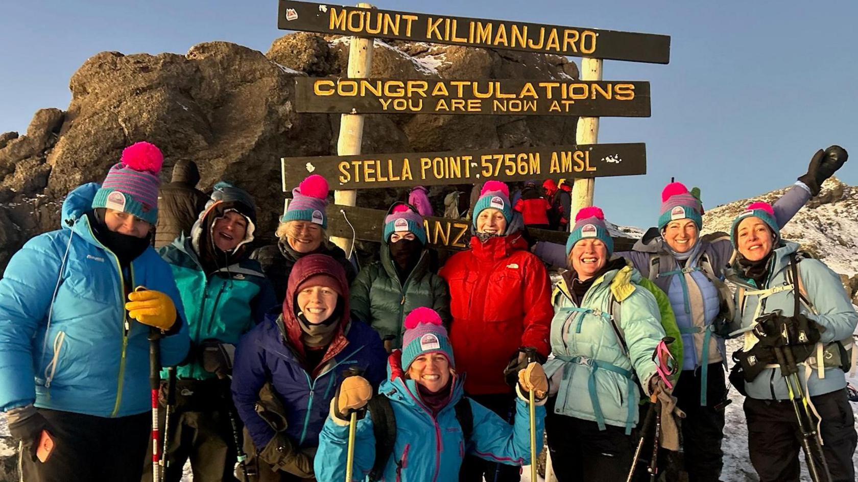 Ten women wearing hiking gear and blue and pink bobble hats pose in front of a sign saying "congratulations, you are now at Mount Kilimanjaro".