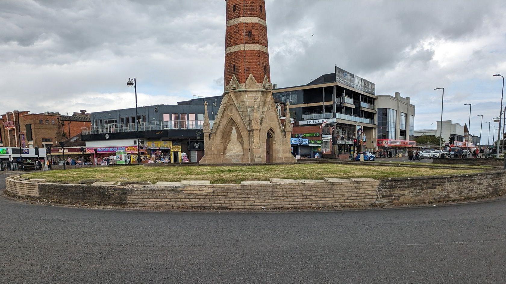 A landscape street view of a circular raised lawn surrounded by a low wall on a tarmac road. The wall is discoloured on the left and right with the yellow/beige stone bricks missing. In the middle is a narrow red brick building shaped like a tower with the base made of stone and featuring gothic edifices. In the background are retail units and commercial buildings