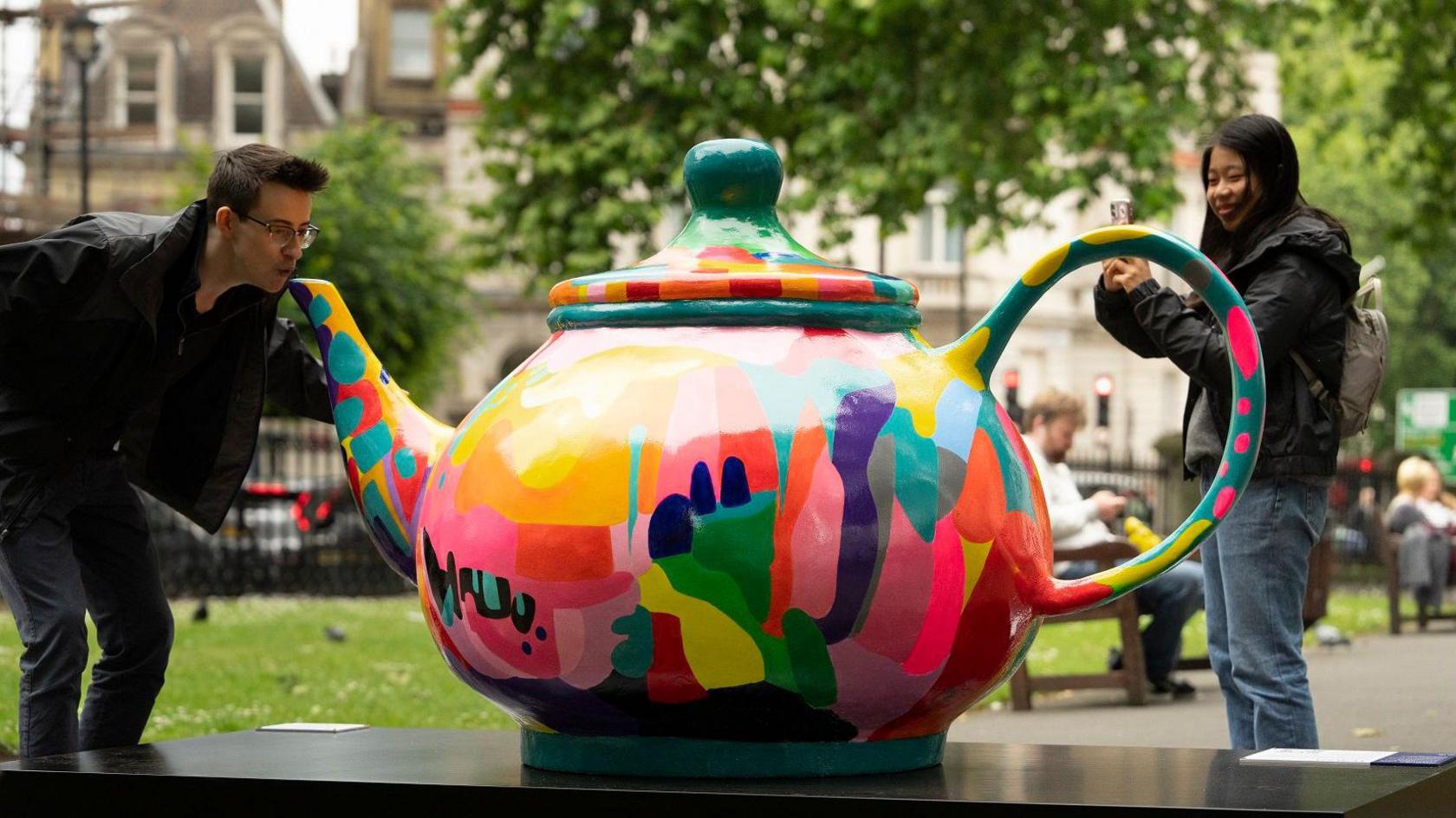 A man pretends to drink from the spout of a giant teapot sculpture whilst someone nearby takes a photo