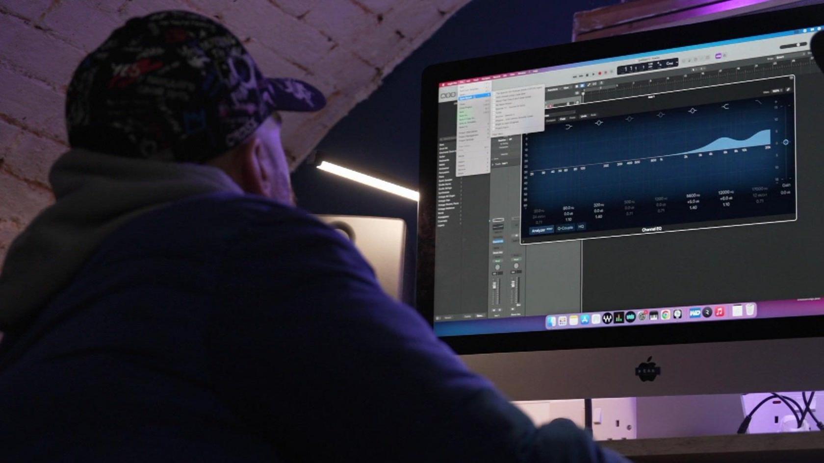 JT sits at a computer with his back to the camera. He is wearing a black cap and a blue puffer jacket. On the screen is music production software. He has a plug-in on the screen which he is using to changing the EQ of a track.