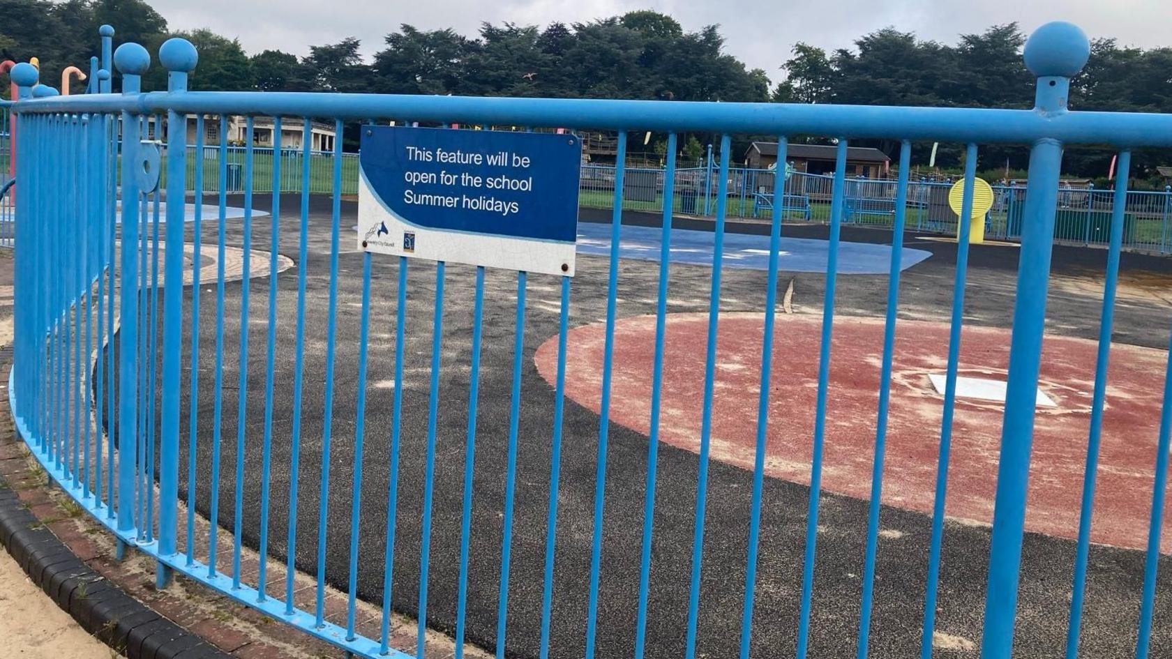 A sign hangs on the railings at the splash pad feature at the park. It reads "this feature will be open for the school summer holidays".