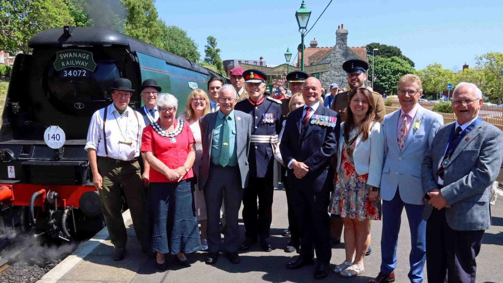 A group of people including a woman in mayoral chains and two men in military uniforms with medals standing on a station platform with a steam locomotive in the background