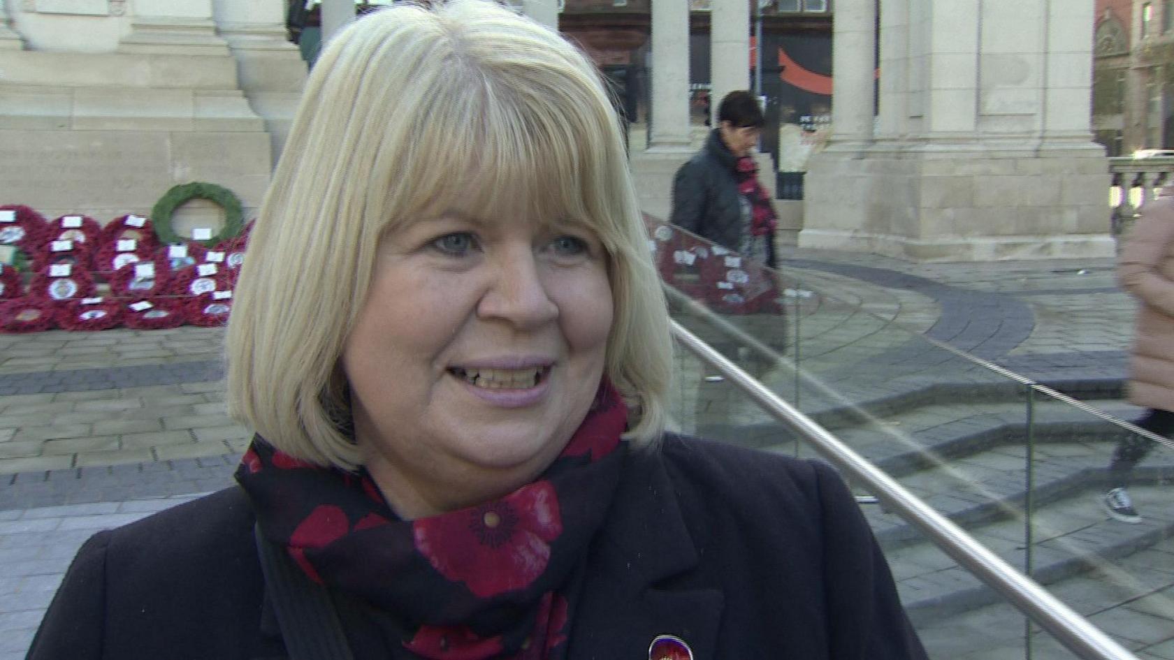 Carol has a blond bob. She's wearing a scarf with a floral design and a jacket. She's standing in the grounds of city hall with wreaths in the background.