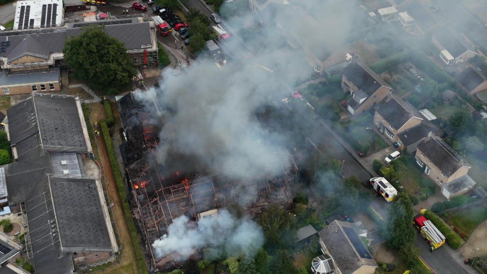 The frame of a building with flames inside and smoke rising above. This is an aerial shot, with fire engines and other emergency service vehicles gathered on streets surrounding the hospital site.