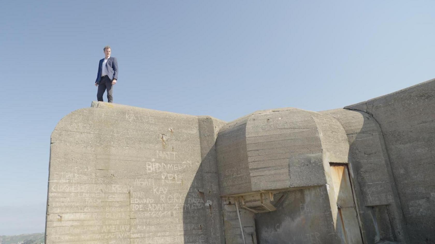 Dan Snow stands on top of a WW2 bunker, with the sea behind him.
