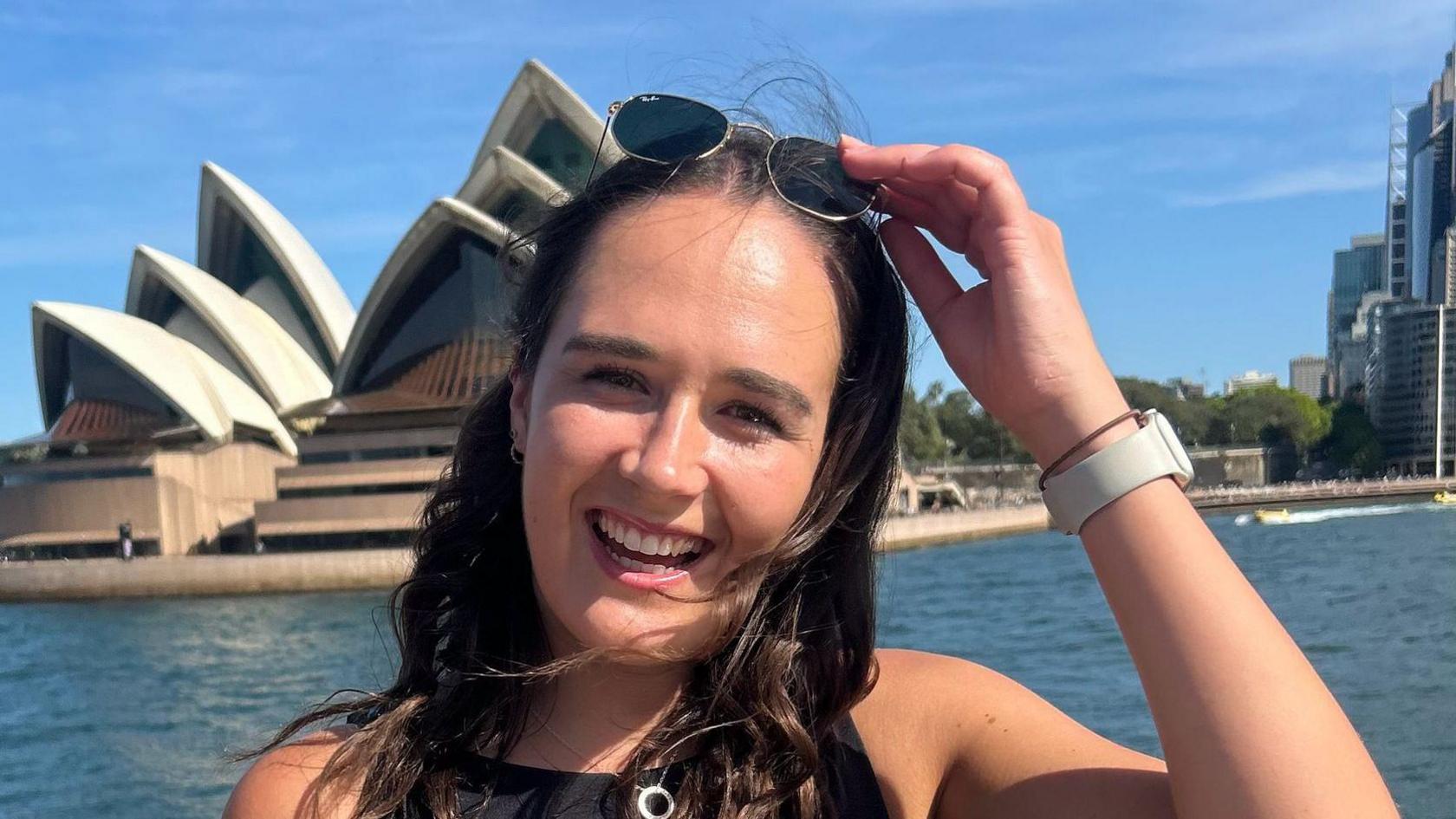 Boireannach le falt dorcha agus speuclairean-grèine air mullach a cinn, air beulaibh Taigh-opera Sydney.