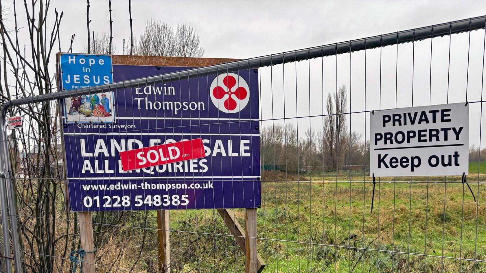 A metal fence with a sign that reads land for sale, sold. There is overgrown grass and bushes behind the fence. Another sign reads 'Private Property, keep out'. It is a wet day. 