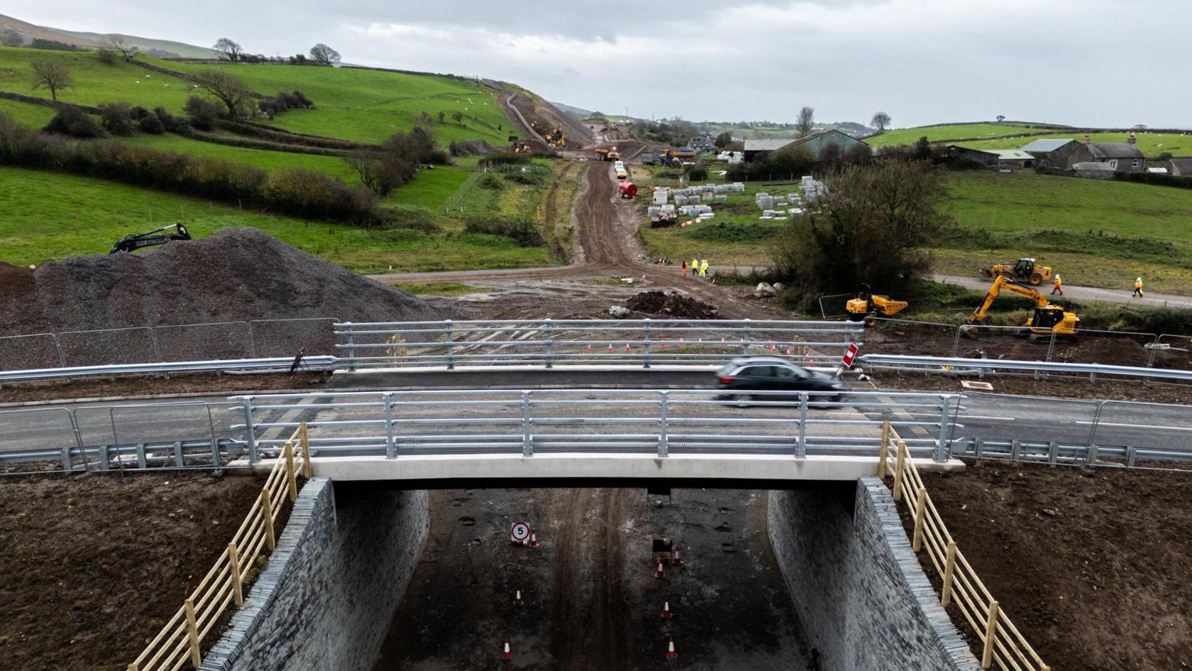 The new bridge with a car in motion travelling on it. The bridge is over an unfinished stretch of road, which looks like a dirt track, with machinery and workers in high viz working on the site.