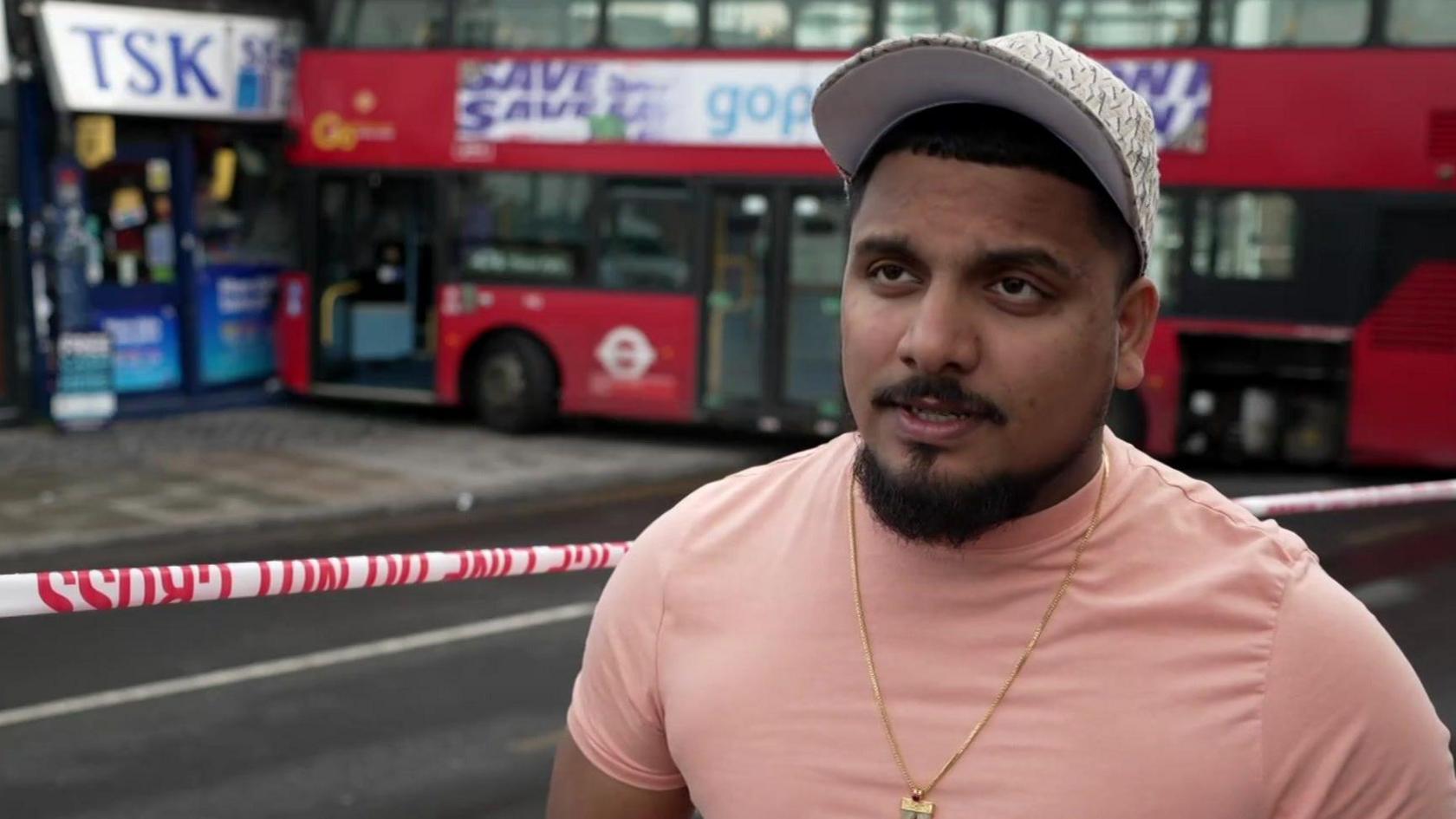 A man in a peach t-shirt and a white cap stands in front a bus that has crashed into the front of a shop in a suburban high street.