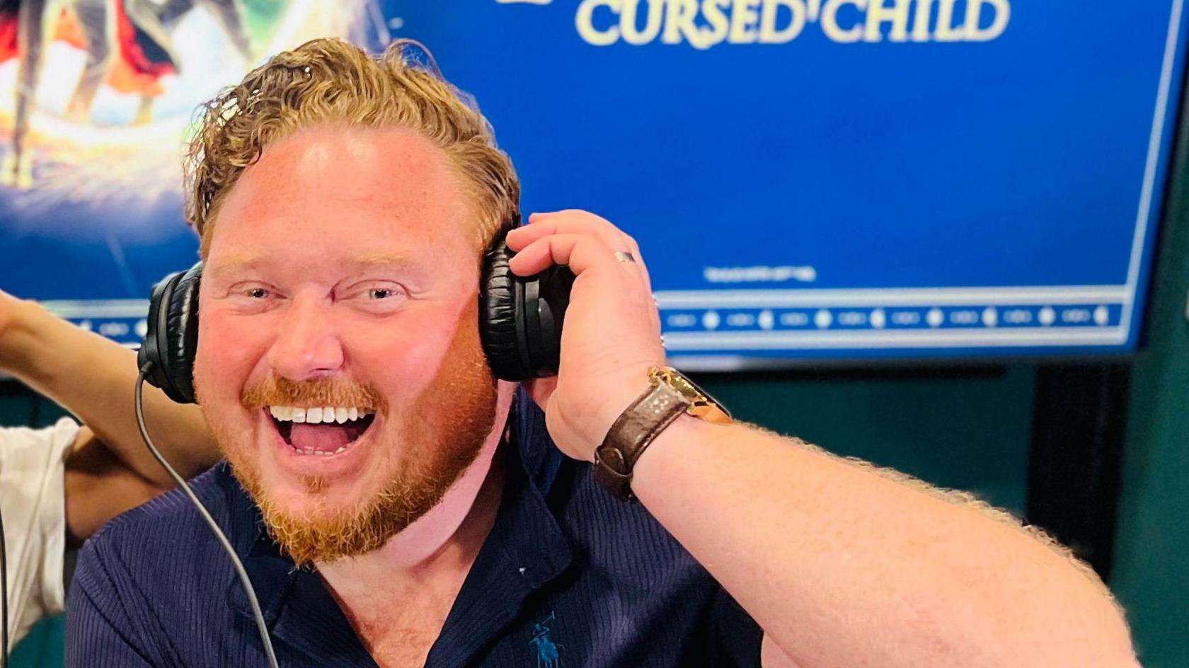 A man with a beard wearing black headphones.
He is a big smile on his face and is holding onto the right side of the headphone.
He is wearing a navy shirt