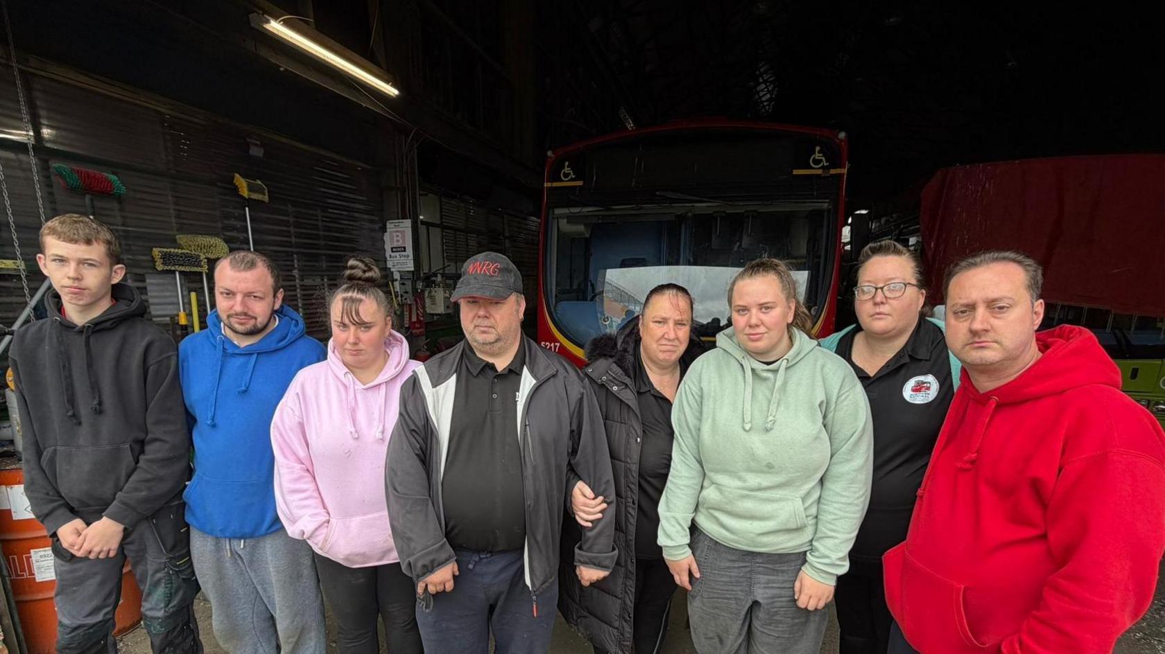 Eight volunteers from the group, including chairman Craig Smith, have lined at the depot. They have serious expressions on their faces. There is a red Go North east bus behind them.