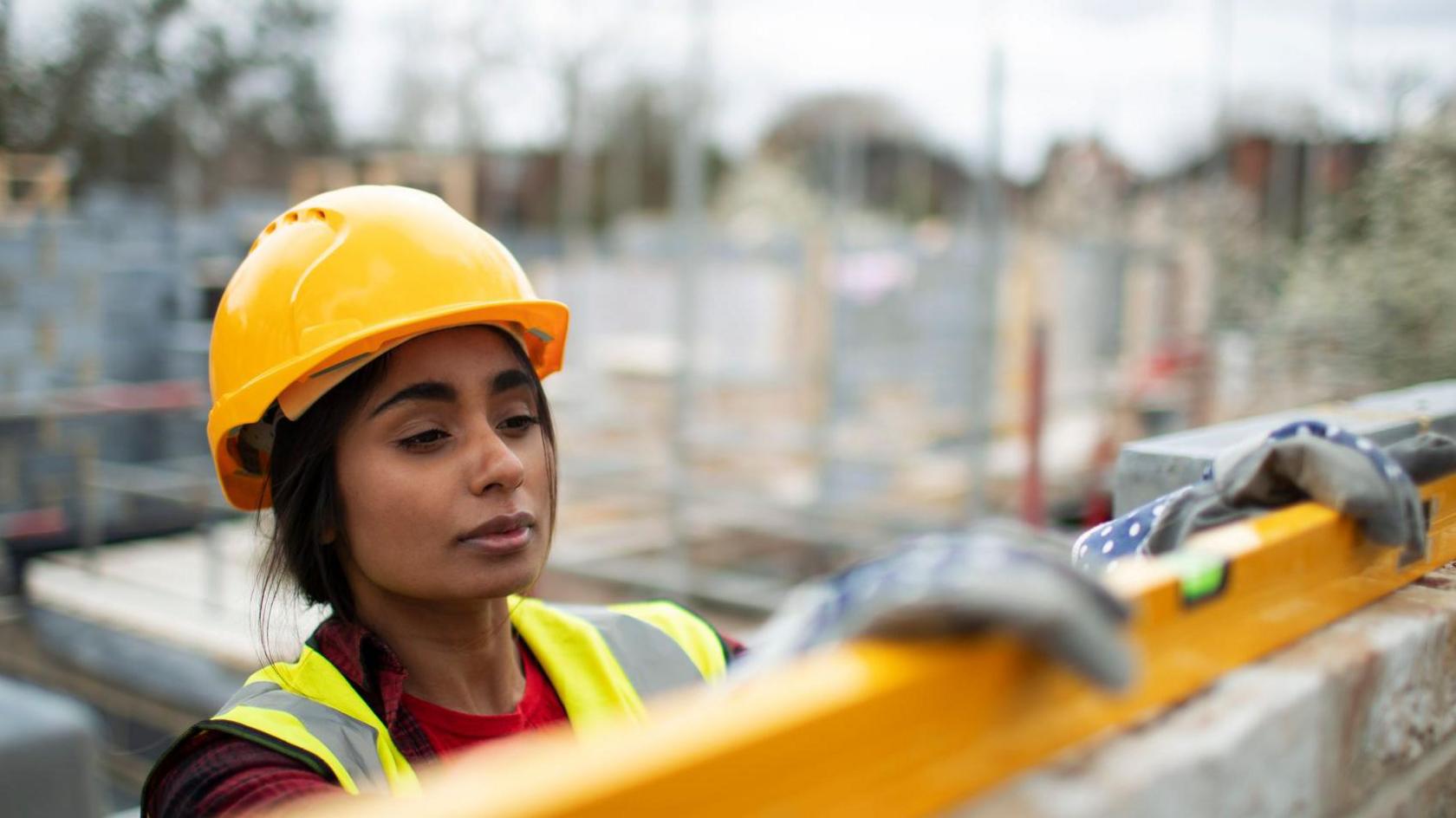 South Asian woman in hard hat and hi viz uses a spirit level on a brick wall. She is outside on a building site.