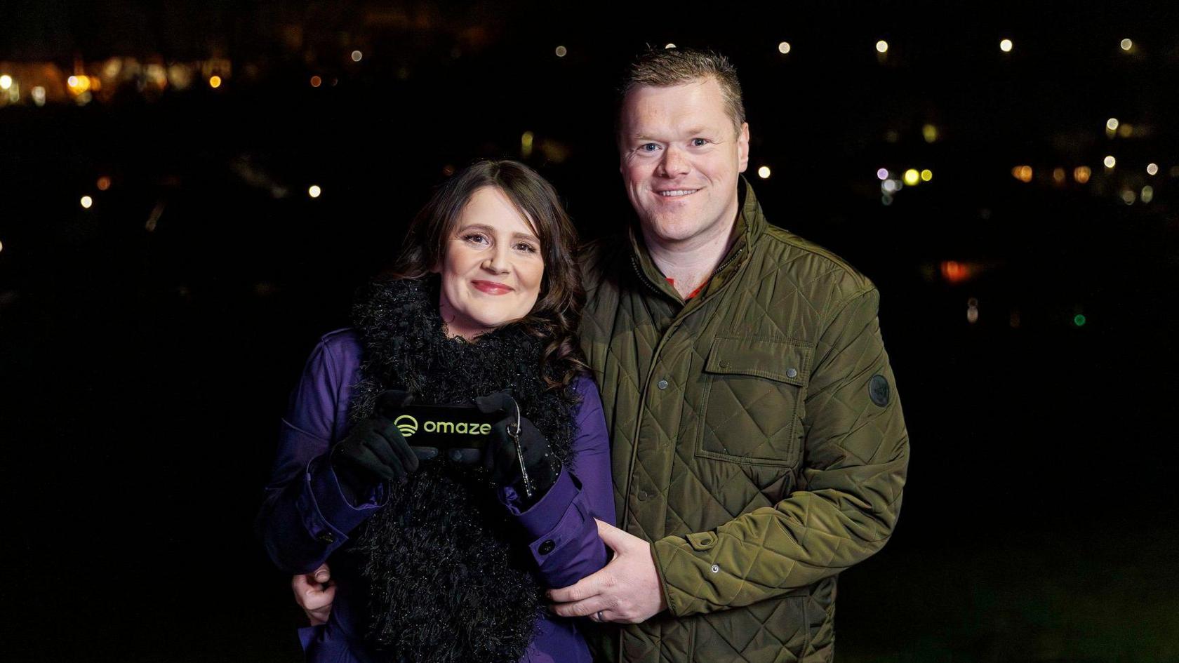 Vicky and her husband Dale standing outside at night with blurry lights in the background. She wears a purple raincoat and large black scarf, and holds a small black pouch with the word Omaze on it. Dale wears a green quilted jacket and has his arm round Vicky. He has cropped light brown hair, she has long wavy dark brown hair.