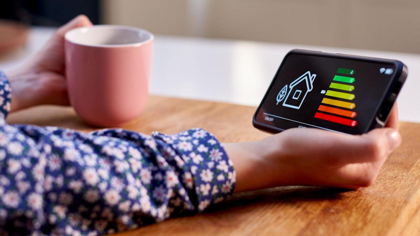 Close up of a woman holding a smart energy meter on a brown kitchen table. 

She is wearing a blue shirt with pink flowers on it and she is holding a a pink mug on her left hand and a black smart energy meter on her right hand that has a sketch of a white house and then varying levels of energy in green, yellow, orange and red lines. 