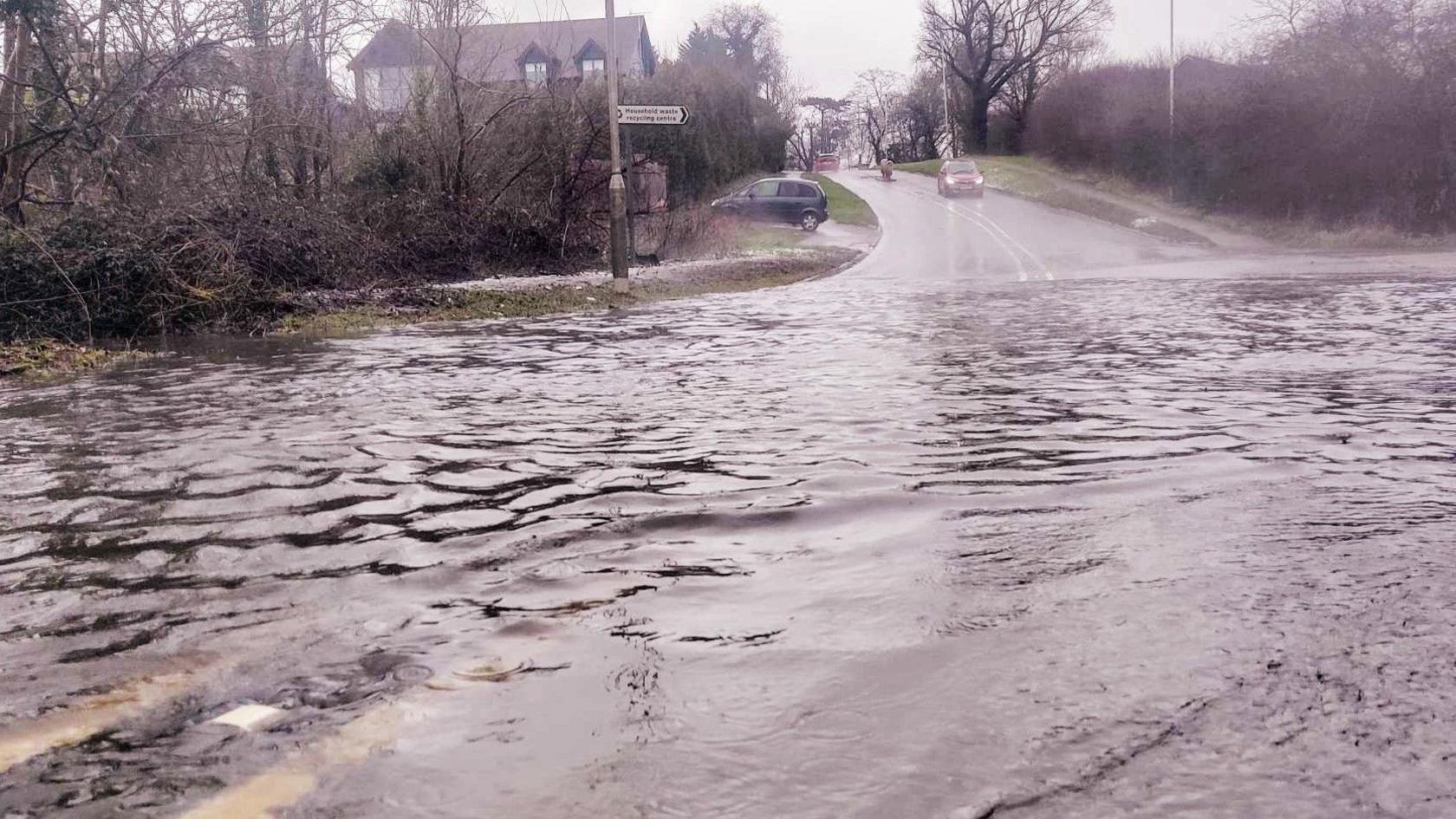 A flooded Allum Lane with vehicles in the background driving down the hill towards the water. The road is lined with trees and there are houses in the background.