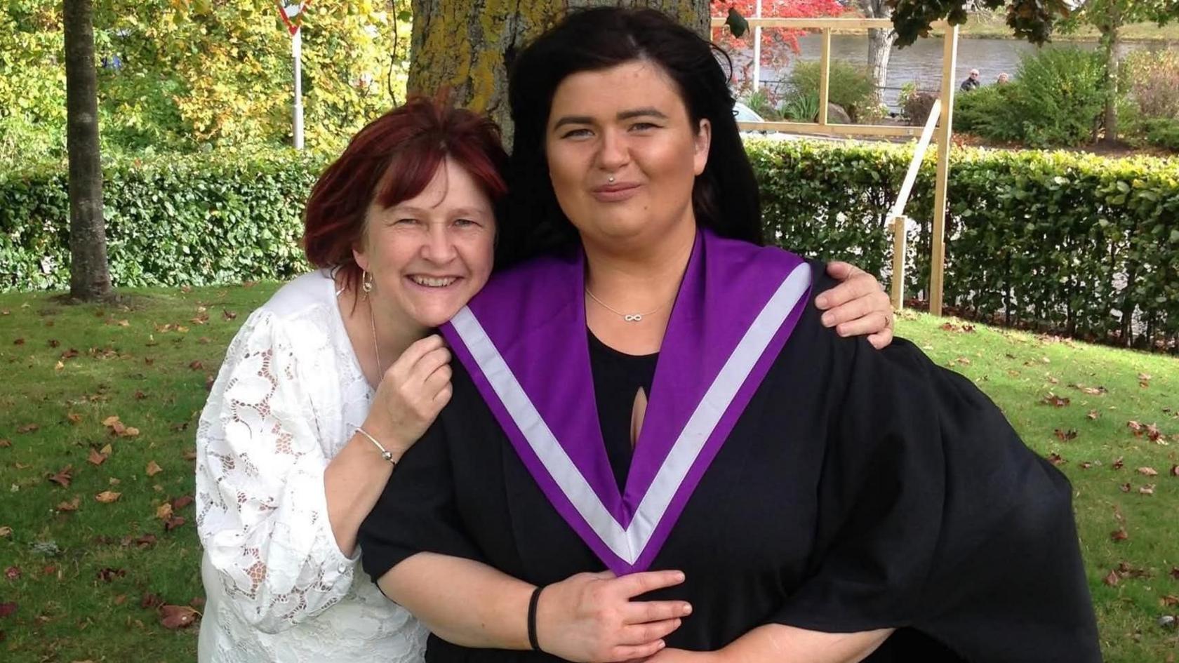 A woman dressed in a graduation gown and with dark hair smiles for the camera with an older woman standing next to her. The older woman is wearing a white top and has her arms around the younger woman. She is also smiling.