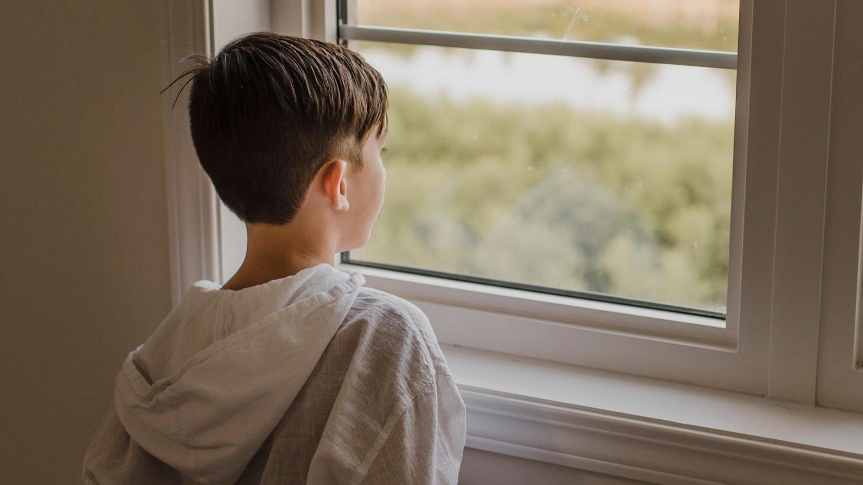 Young boy looking out of a window, wearing a white hoodie. The hood is hanging down his back. The boy is viewed from the back of his head, with the window in view.