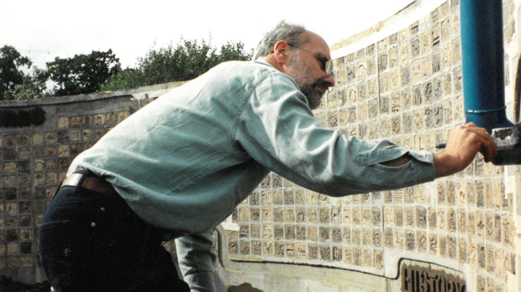 Christopher White using a paintbrush on some of the tiles on the wall. He is concentrating on his task and wears glasses, a blue shirt and black jeans