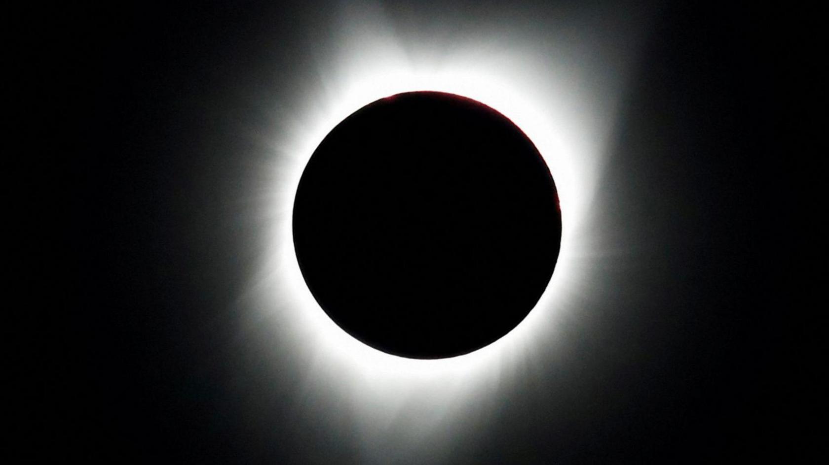 A total solar eclipse with the white rays of the sun peeking out behind the moon which is obscuring it, with a dark black sky behind it.