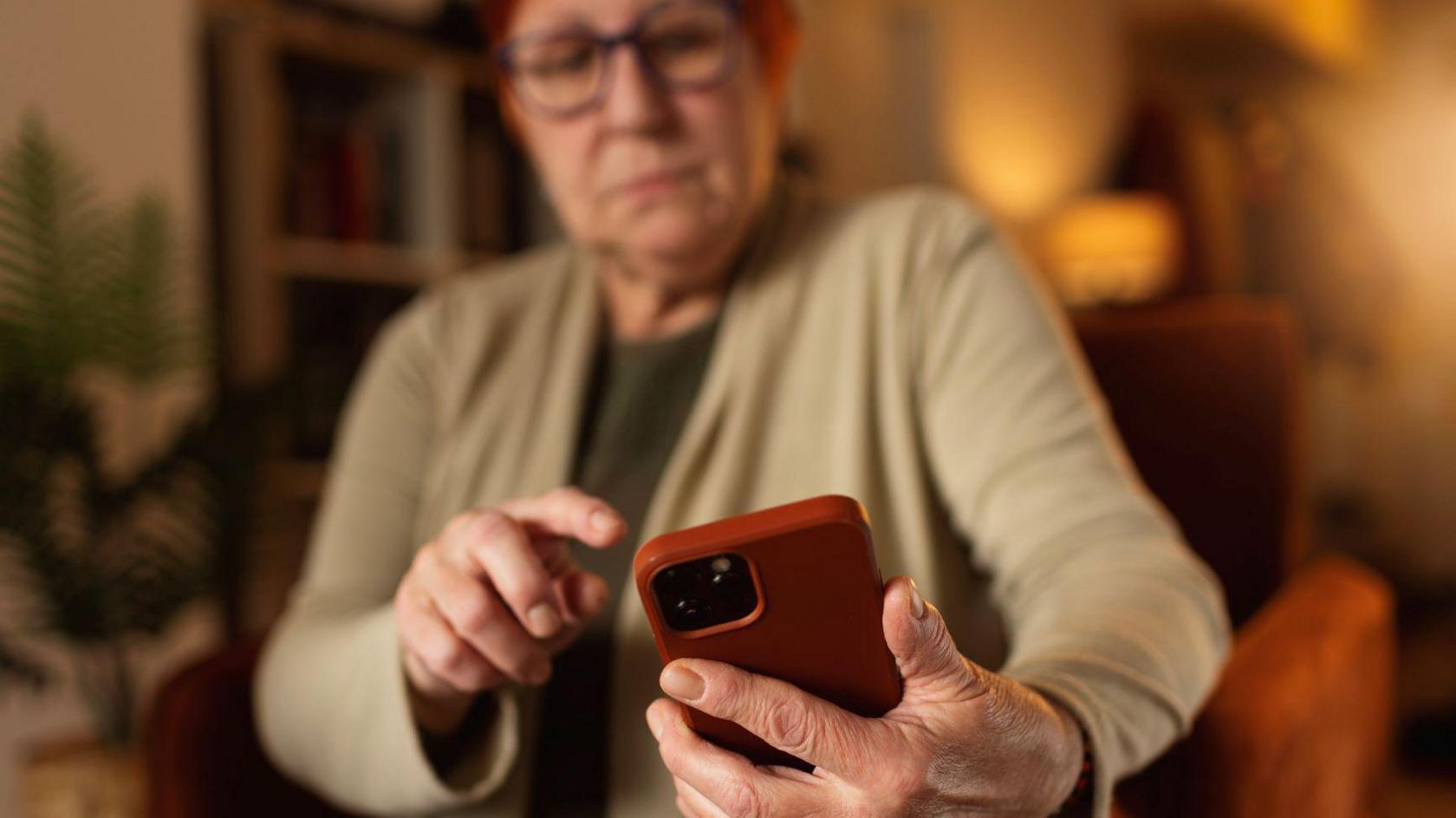 A woman holds a smartphone. She has short dyed red hair and glasses, and she is wearing a beige cardigan. The smartphone is red.