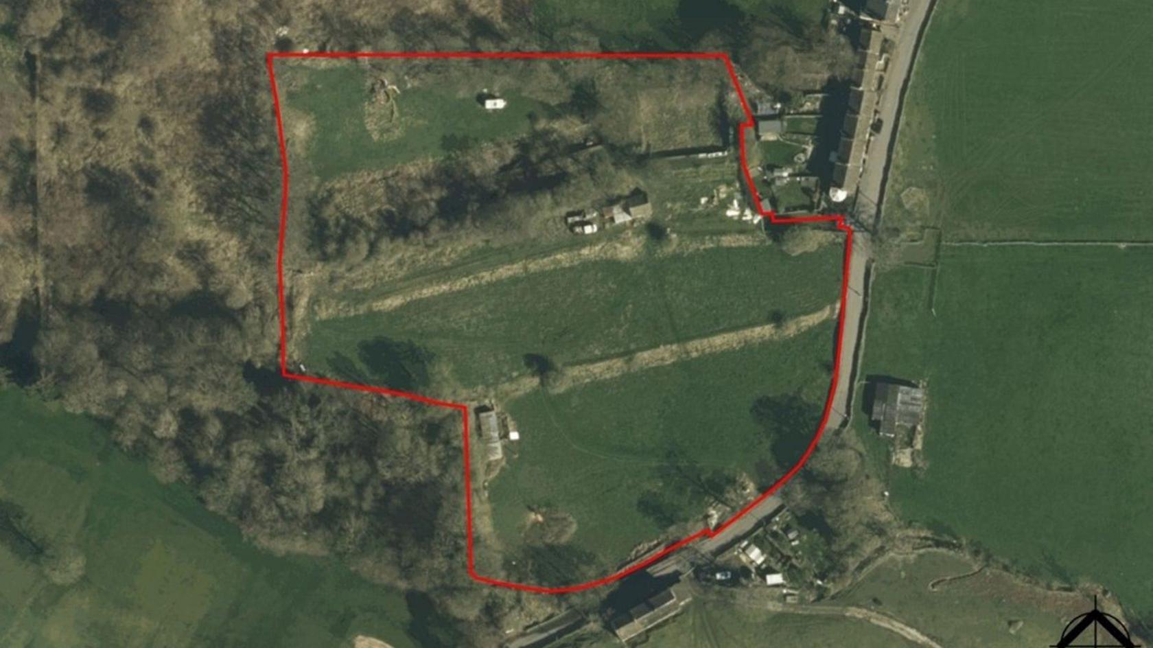 Aerial photograph of a semi-rural property outlined in red. The plot includes grassy fields, scattered trees and several buildings. A road runs along the right edge, with additional structures nearby. Wooded areas border the left side.