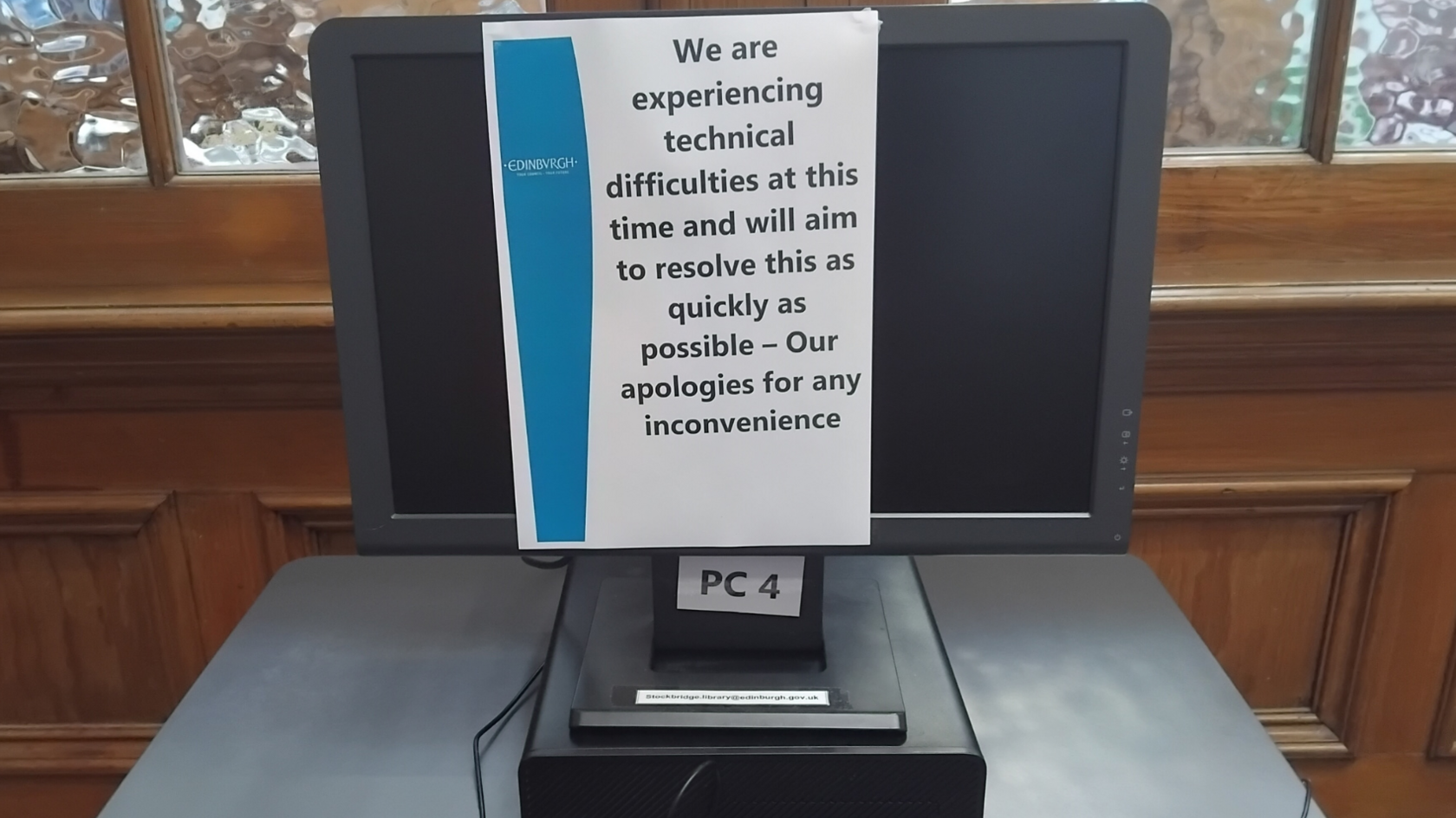 A black desktop PC screen at a library in Edinburgh which has been turned off and has an A4 notice taped to it. The notice reads: "We are experiencing technical difficulties at this time and will aim to resolve this as quickly as possible - Our apologies for any inconvenience". The screen is on a grey table in front of a wooden background with frosted glass windows.