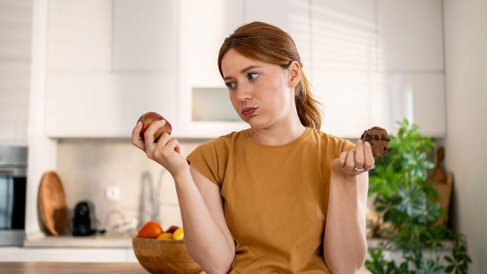Young woman holding an apple and a chocolate cookie, contemplating a healthy food choice in a stylish, modern kitchen