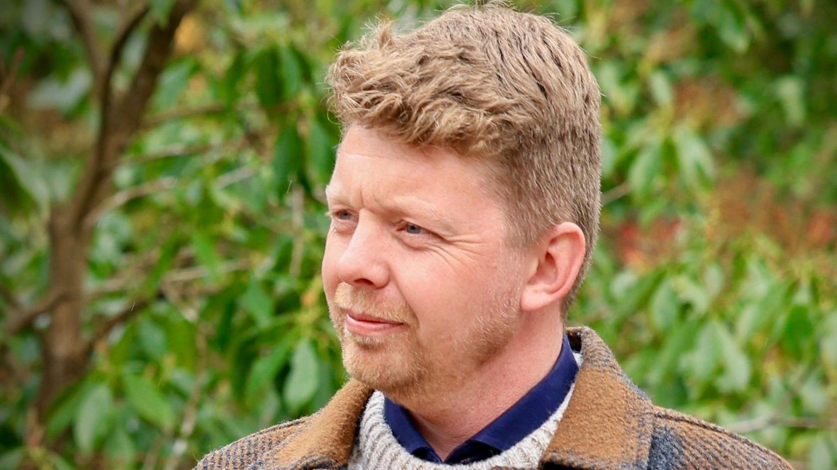 Peter Jones looking to he left with trees in the background. He's wearing a yellow and grey jacket and has blonde hair and beard