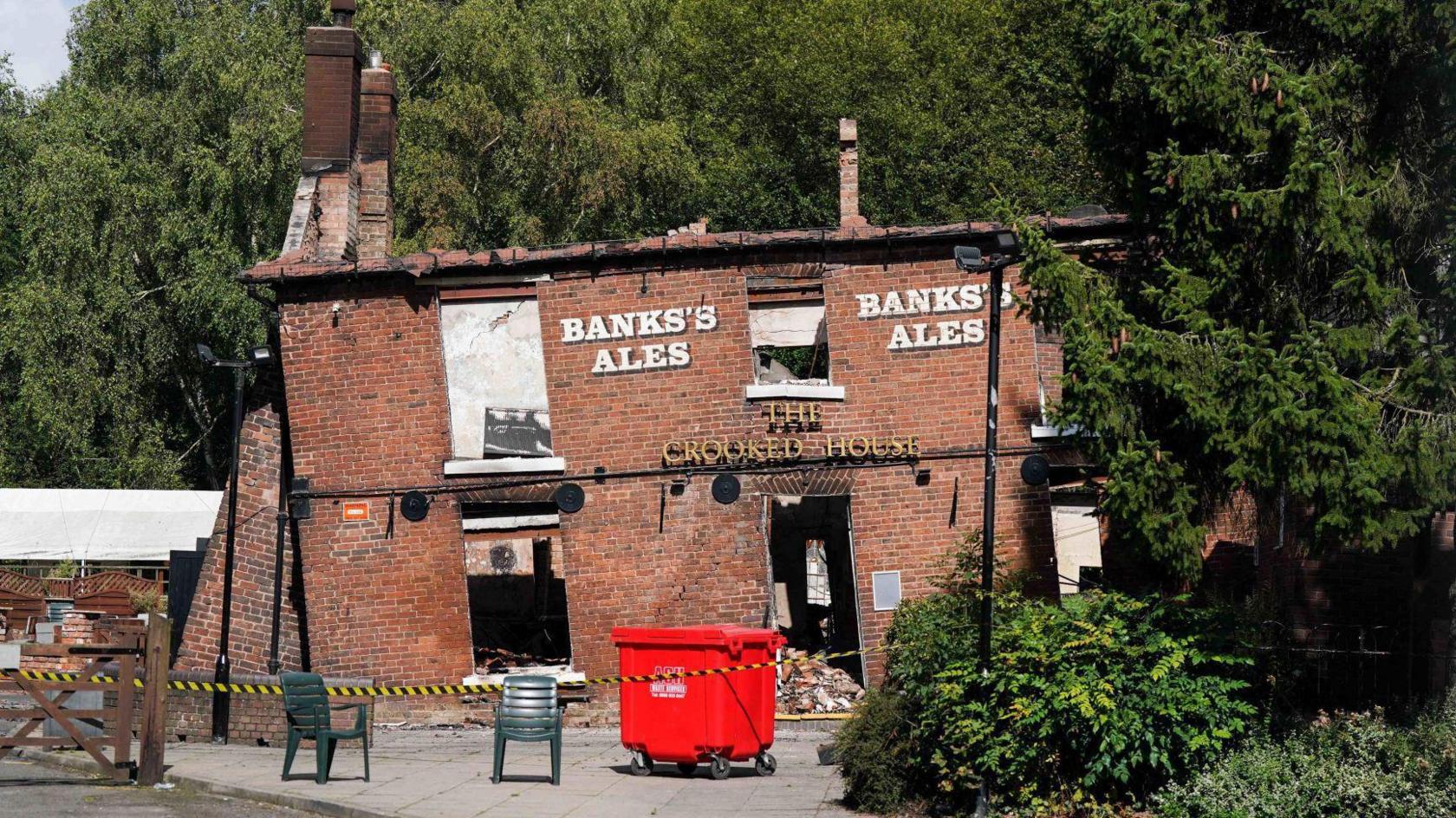 The crooked house, a pub that sits at a slanted  angle. it is a red brick building and is damaged by fire, with tape surrounding it.