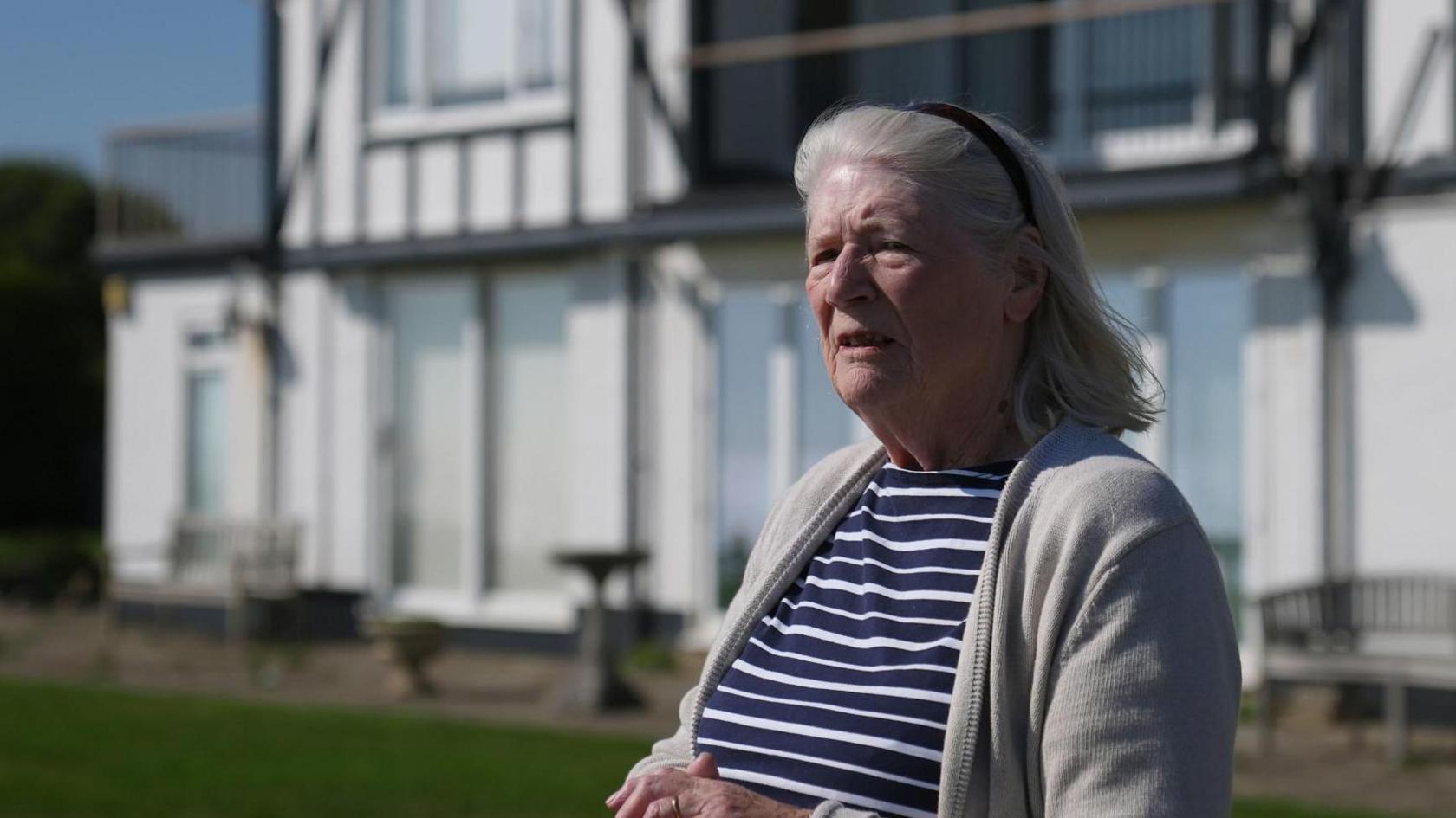 Jean Flick wearing a blue and white striped top and a white cardigan, she is wearing a black headband. She is standing in front of her large white home.