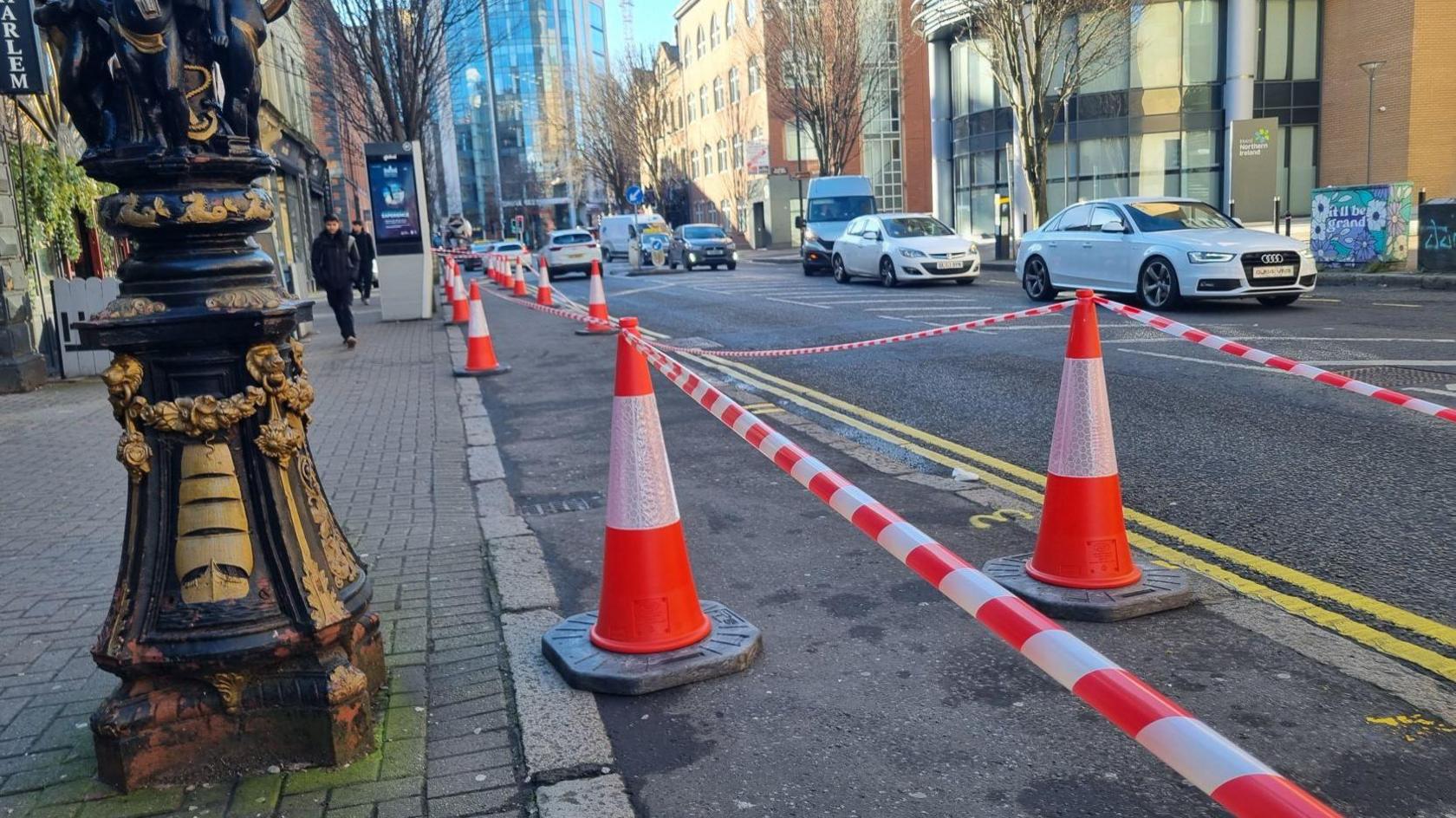 Rows of fluorescent orange cones with tape strung between them on a footpath along Bedford Street in Belfast