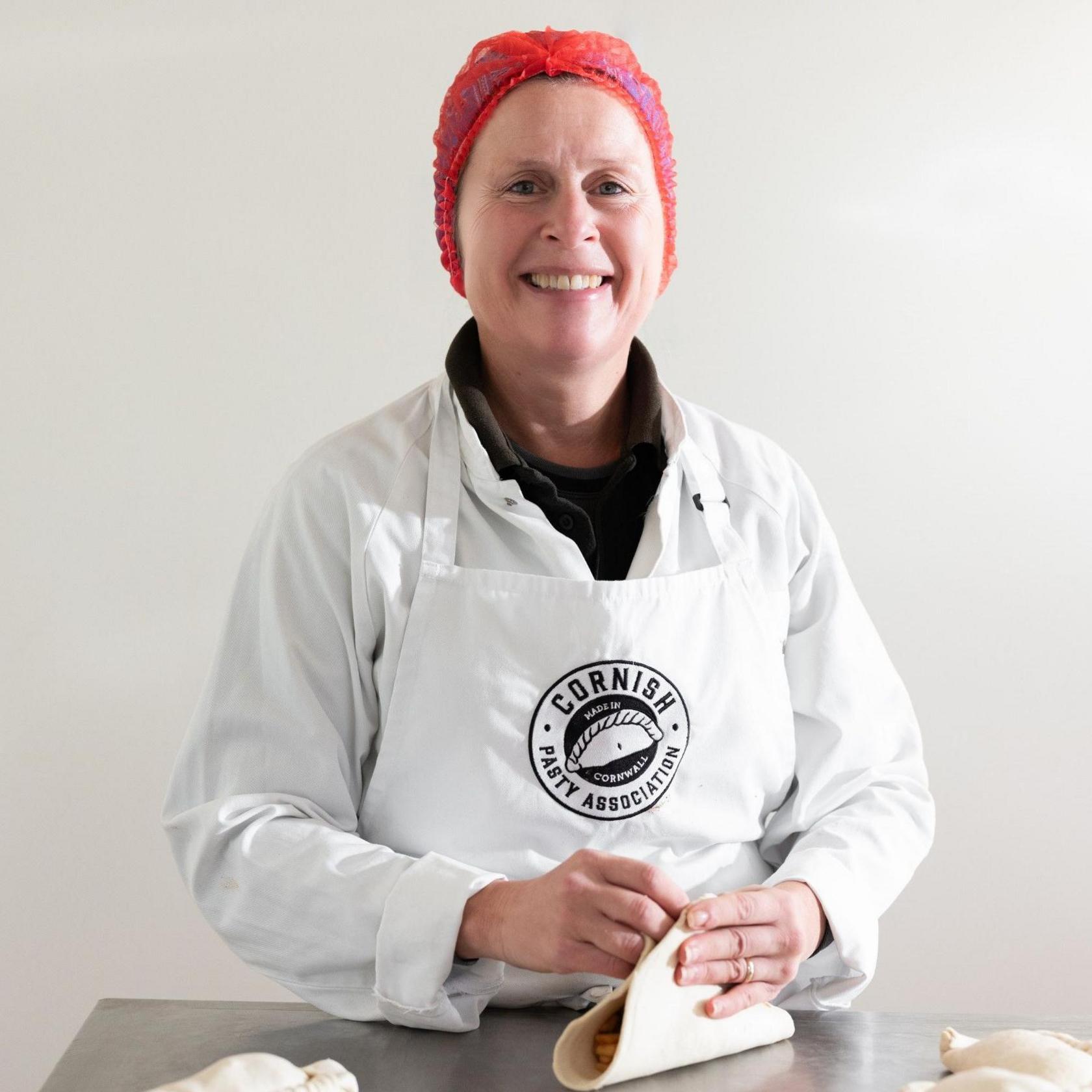 Professional baker Jackie Tonkin of the Cornish Pasty Association  is smiling at the camera as she crimps a pasty. She is wearing a white coat, a white apron with a CPA logo and a red hair net. 