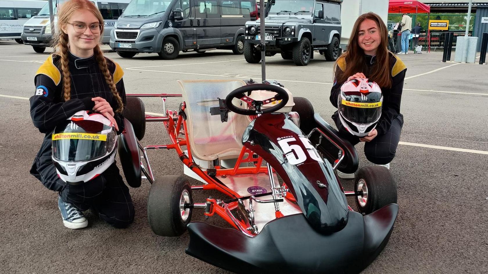 Chloe and Lucy are in racing suits are kneeling on either side of a go-kart with the number 5 on its front. They are holding helmets and posing for a photo in what appears to be a parking area with several vehicles and tents in the background
