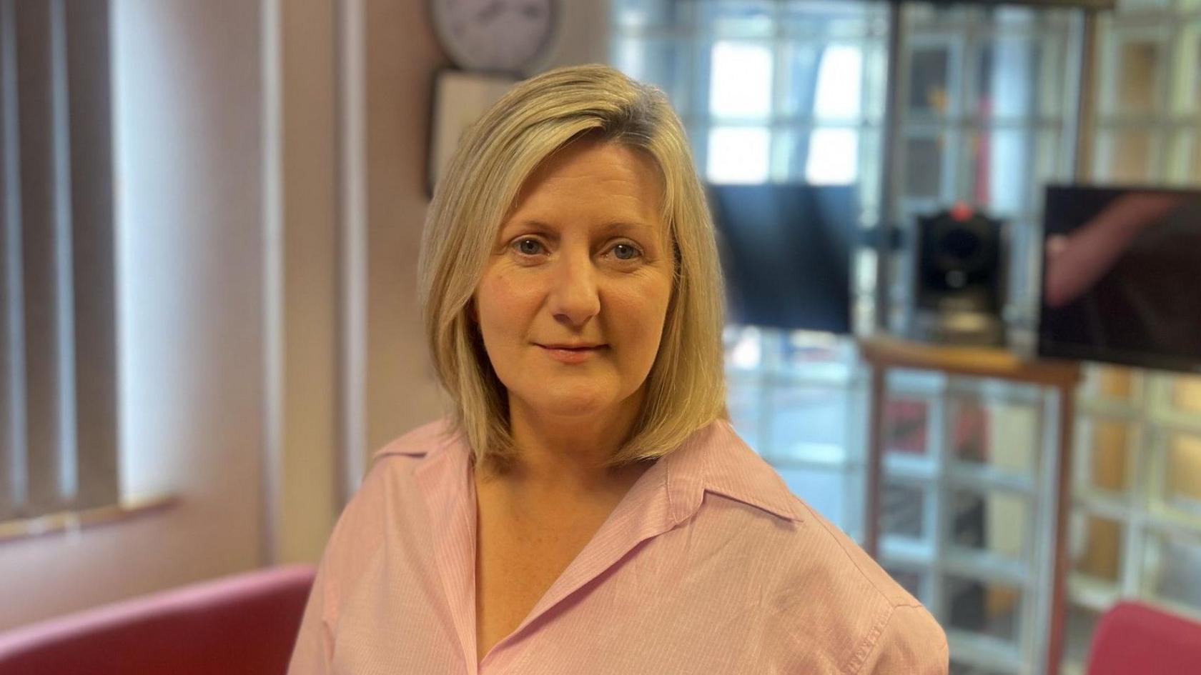 A blonde hair woman wears a pink shirt. Behind her left shoulder are two black tv screen mounted on a wall. Below the TV's are tow red sofas. Over her right shoulder is a closed white window blind.
