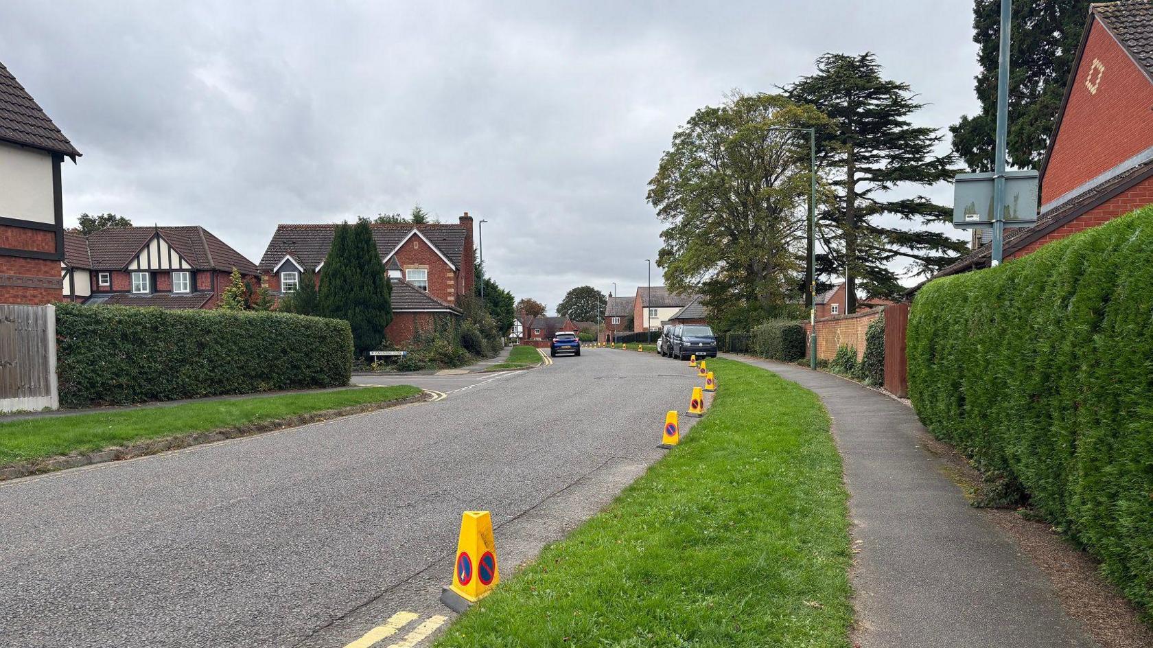 A stretch of road on a residential street with houses and pavements either side. There are yellow traffic cones on the right hand side, between stretches of double yellow lines.