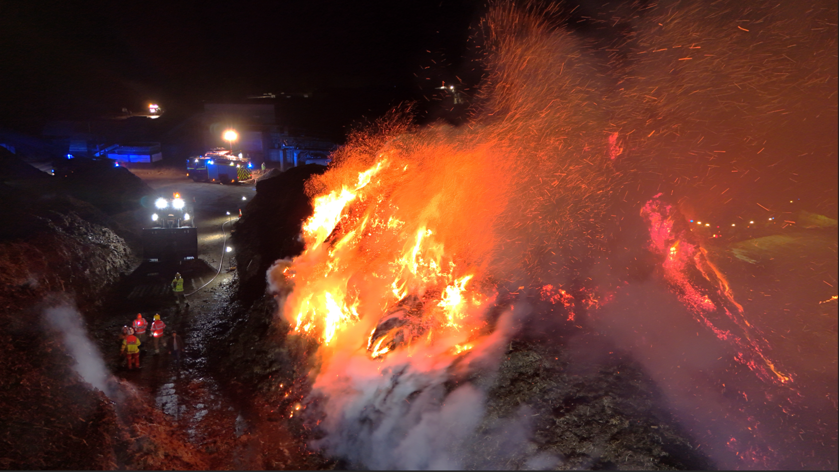 Two large piles of bark are shown with four firefighters standing between them. the left pile is smouldering but the right is fully ablaze but rest of the site is mostly in darkness other the lights of some vehicles.