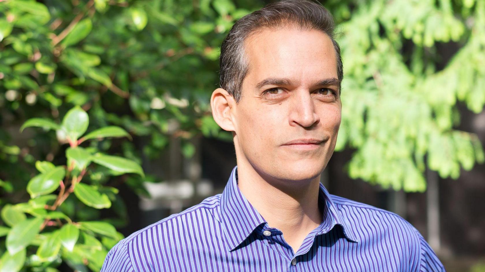 Prof Will Dixon is standing in front of a background of greenery. He is wearing a blue stiped button down shirt